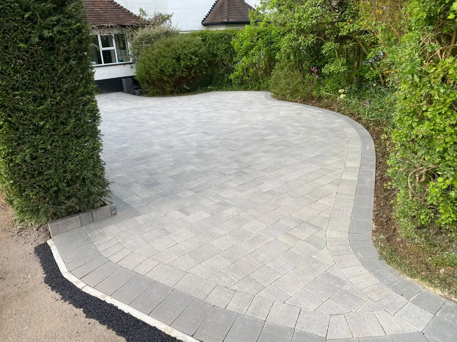 Pale grey block paving with a curved grey border sweeping round a hedge and tree